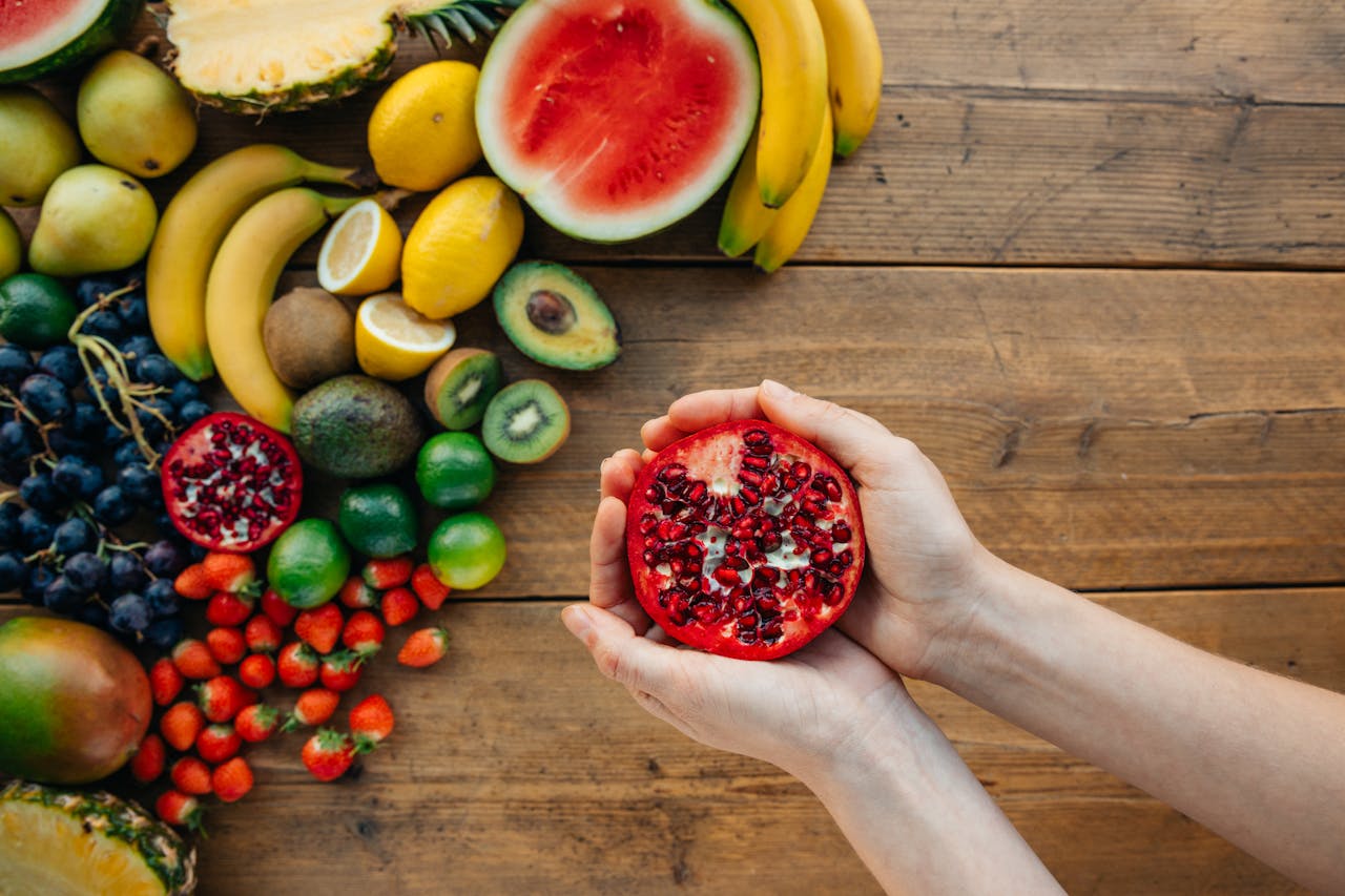 Hands holding pomegranate with a vibrant assortment of fresh fruits on a wooden table.