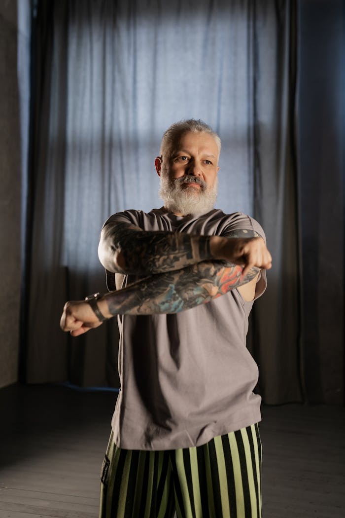 A tattooed senior man performing a stretch indoors, promoting fitness and wellness.